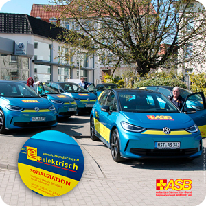 Es stehen 6 t&uuml;rkiesblaue VW ID.3 vor dem VW Autohaus, mit gelb-roter ASB-Beklebung. Hinter jeweils einem Fahrzeug stehen der F&uuml;hrparkmanager, Herr H&auml;gemann-Schenke, die Pflegedienstleiterin Frau Femmer und eine Pflegekraft, Frau Woyzek