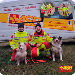 Ein Mann mit Hund und eine Frau mit Hunde, knien vor einem Transprter mit der Aufschrift ASB rettungshundestaffel. Beide tragen Einsatzkleidung. Dicke gelb rote Winterjacken mit Reflexionsstreifen.