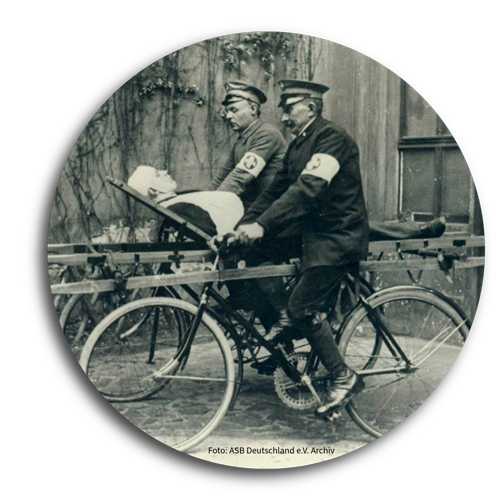 Ein schwarz-wei&szlig; Archivbild. Darauf zu sehen zwei Samariter, die mit dem Trageliegefahrrad eine verletzte Person transportieren. Beide Fahrer tragen eine Armbinde mit dem Samariterkreuz drauf.
