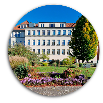 Im Hintergrund das mehrst&ouml;ckige Gesch&auml;ftsgeb&auml;ude des ASB, rechts davor ein gro&szlig;er gr&uuml;ner Baum und im Vordergrund der Innenhof mit einer gr&uuml;nen wiese und verschiedenen Beeten